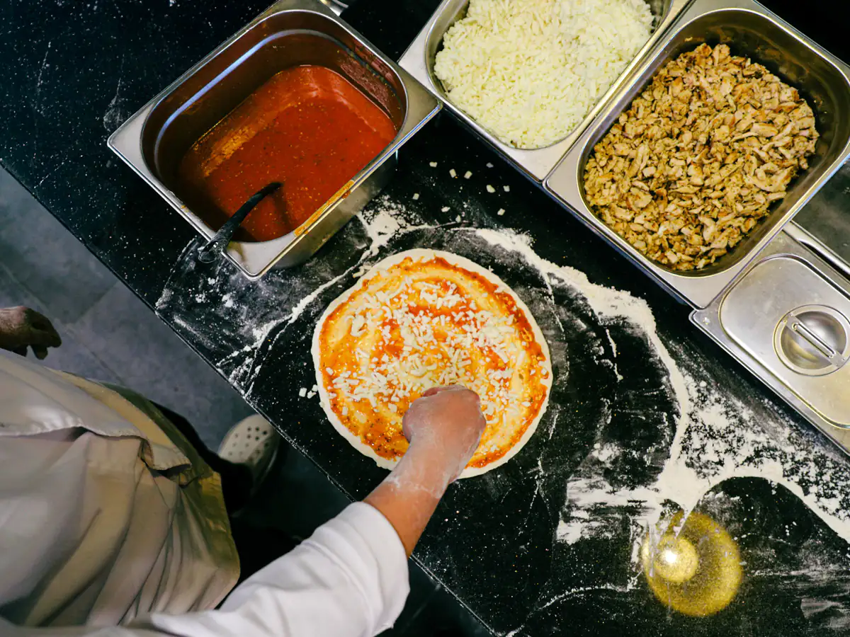 Pizzaiolo montando a pizza sobre bancada enfarinhada, com as cubas de ingredientes ao lado